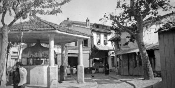 Η πλατεία το 1935 με τον φακό της Έλλης Παπαδημητρίου. Εκτός από την βρύση και τα δέντρα που κοσμούν την πλατεία, ο σχηματισμένος δρόμος με τους μπαβέδες που ξεκινούσε από την Πόρτα Μαγγιόρε, περνούσε από την πλατεία και οδηγούσε στον οικισμό, 1935, Αρχεί Η πλατεία το 1935 με τον φακό της Έλλης Παπαδημητρίου. Εκτός από την βρύση και τα δέντρα που κοσμούν την πλατεία, ο σχηματισμένος δρόμος με τους μπαβέδες που ξεκινούσε από την Πόρτα Μαγγιόρε, περνούσε από την πλατεία και οδηγούσε στον οικισμό, 1935, Αρχεί